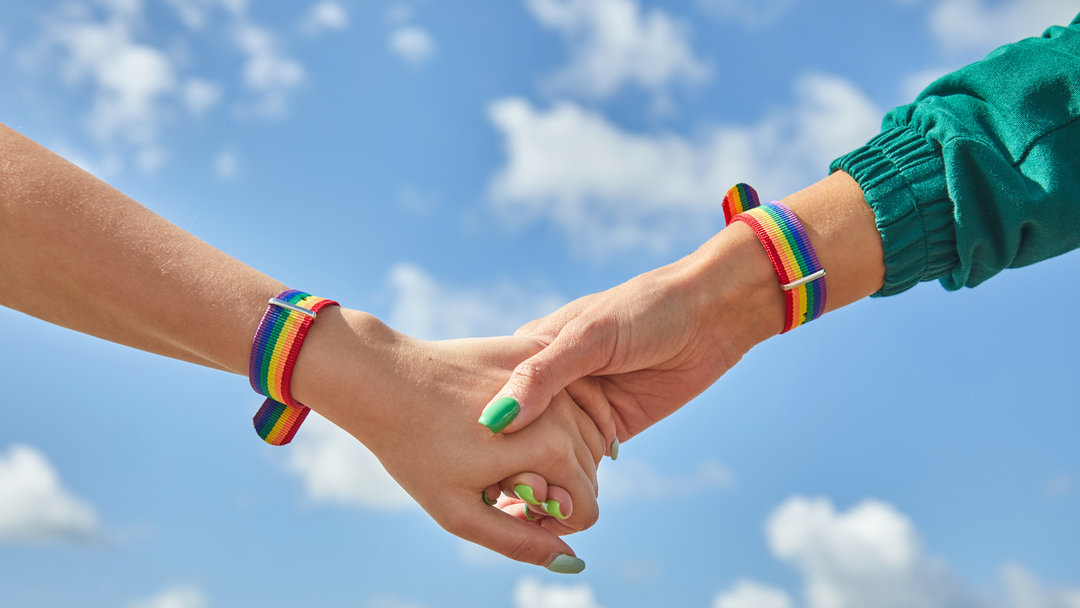 Due donne si tengono per mano con al polso un braccialetto con i colori del Pride